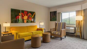 A hotel room with a yellow sofa, a striped armchair, two round ottomans, a large painting of flowers, a standing lamp, and a window with curtains.