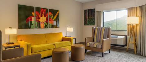 A hotel room with a yellow sofa, a striped armchair, two round ottomans, a large painting of flowers, a standing lamp, and a window with curtains.