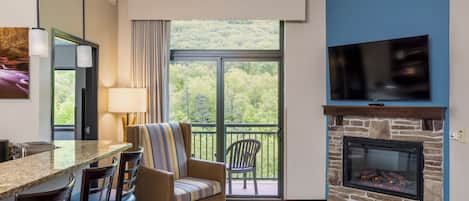 Habitación de hotel con zona de comedor, chimenea, televisión y balcón con vistas a árboles.