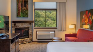 Ein Hotelzimmer mit Kamin aus Stein, einem großen Fenster mit Blick auf Bäume, einem roten Sofa und einem Flachbildfernseher.