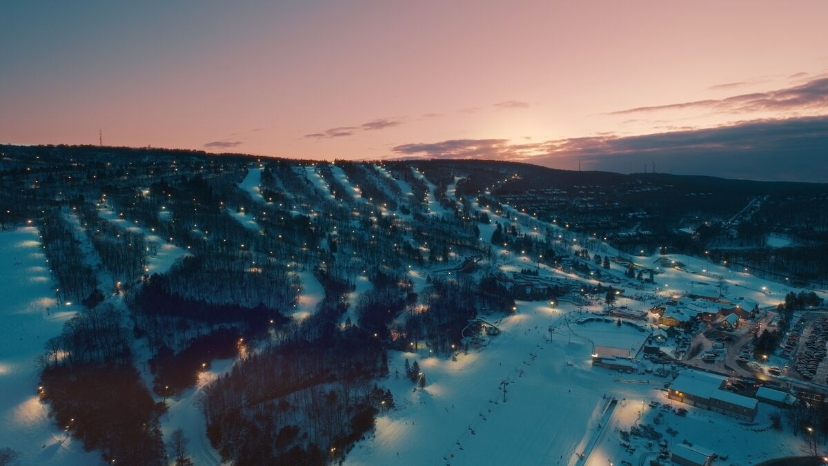 Lumisateen peittämä maisema, jossa näkyy kaupunki hämärän aikaan. Kuvassa on valaistuja rakennuksia ja mutkitteleva tie.