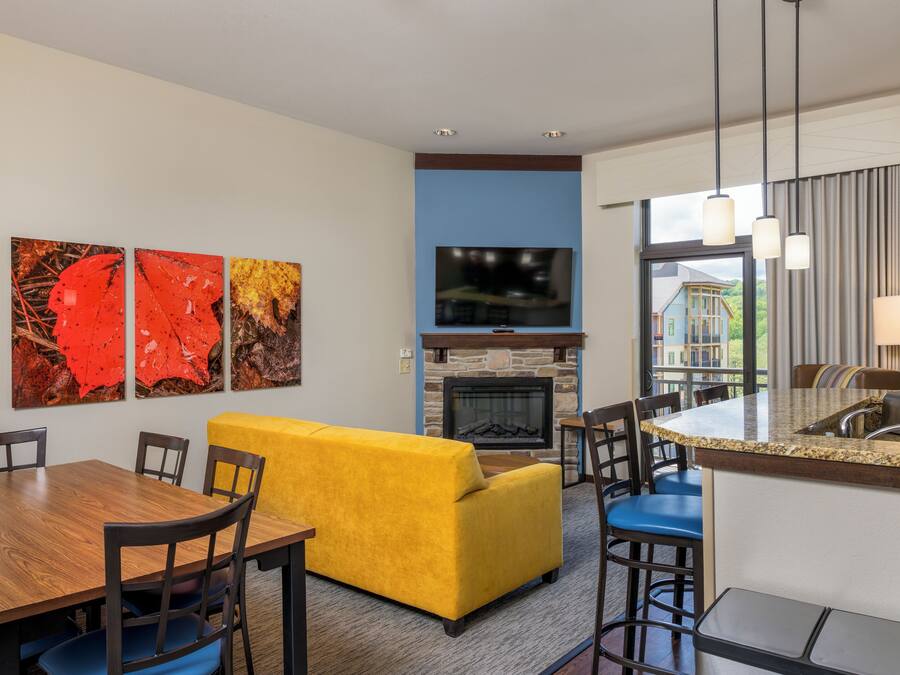 Un salon moderne avec un canapé jaune, une cheminée, un téléviseur et un coin repas comprenant une table et des chaises en bois.