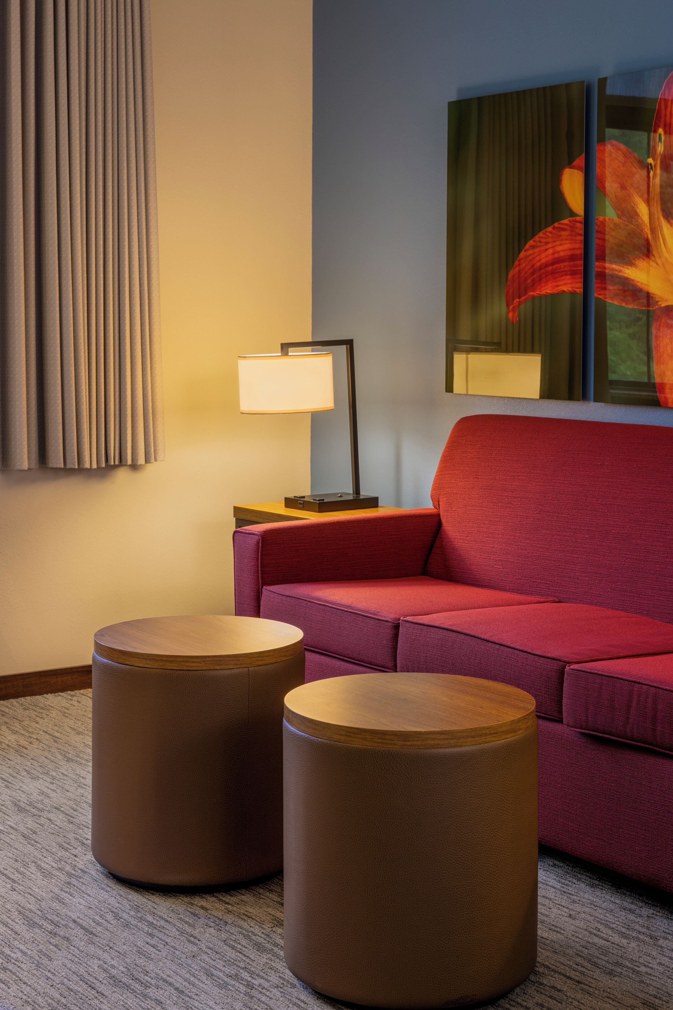 A modern hotel room with a red sofa, two wooden ottomans, a lamp, and a large floral painting on the wall.