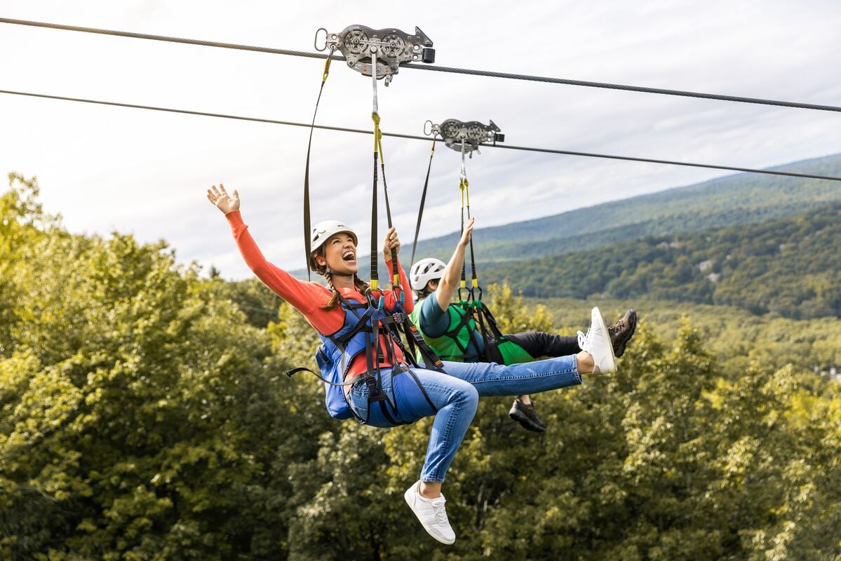Two people suspended in the air on a zip line, surrounded by a forested landscape.