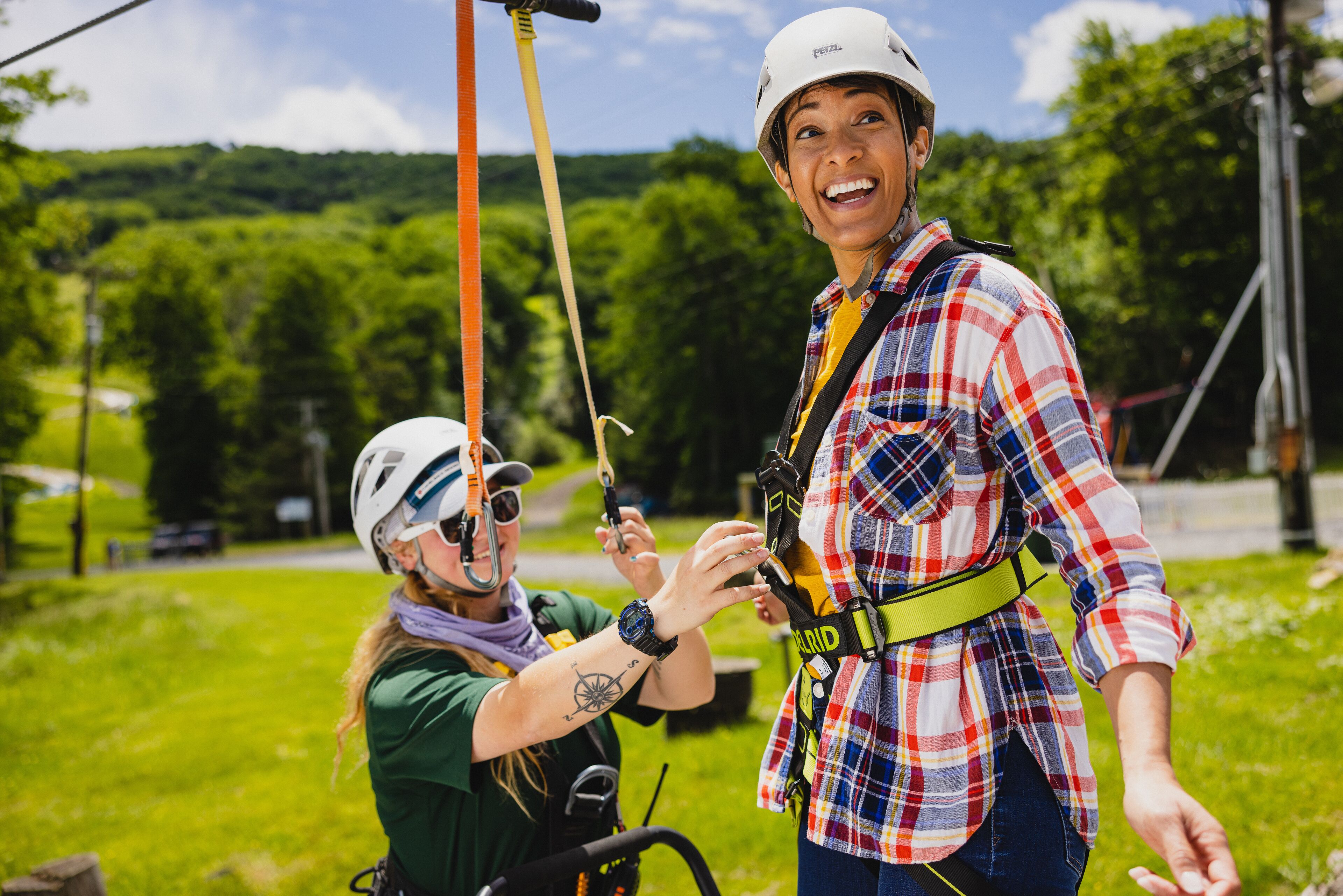 Two individuals in a grassy outdoor setting, one wearing a helmet and harness, the other in a plaid shirt and cap, both appear to be engaged in an activity involving ropes.