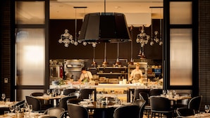 A restaurant with tables set for dining, a kitchen in the background, and hanging light fixtures.
