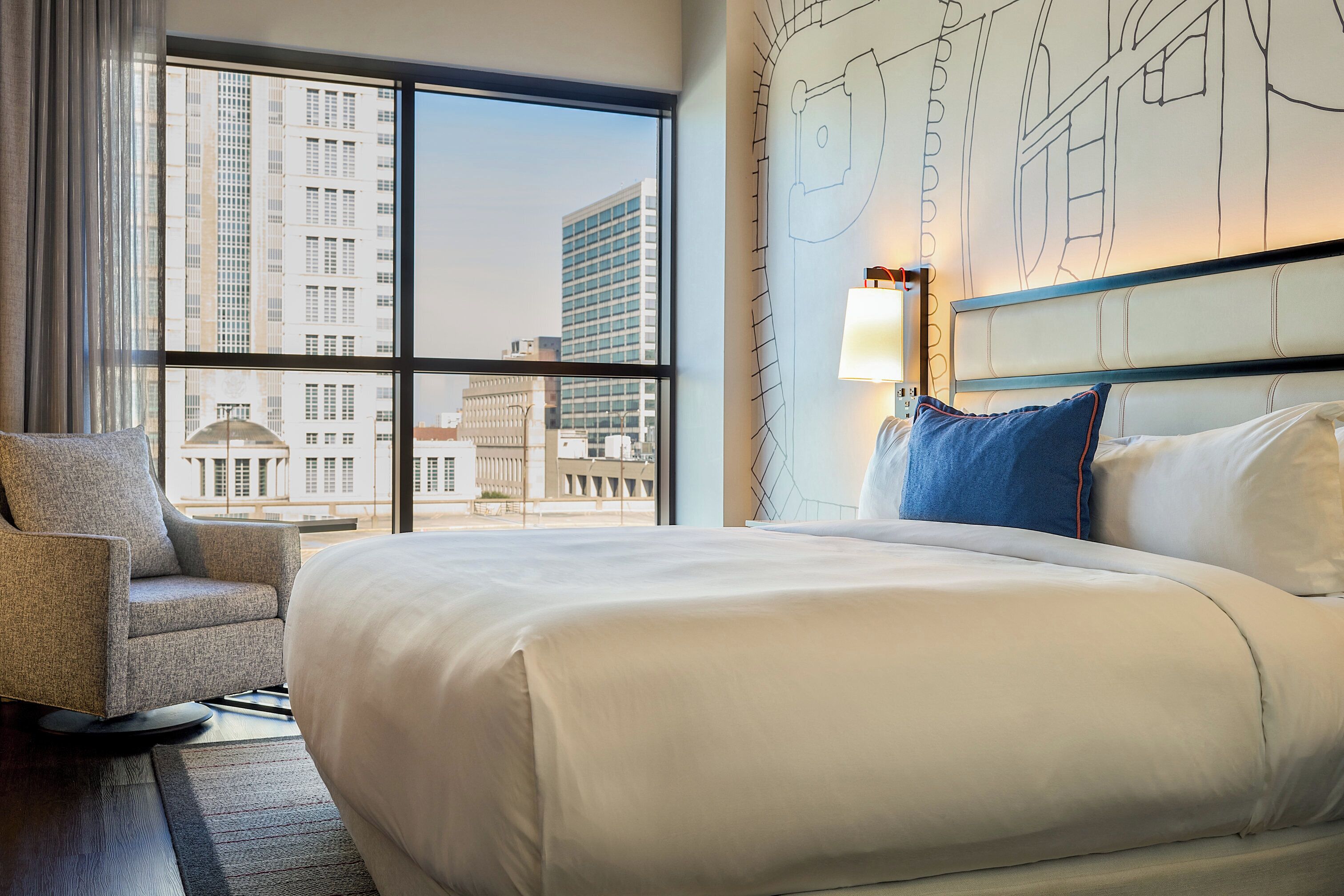 A hotel room with a large bed, a chair, and a view of tall buildings through large windows.