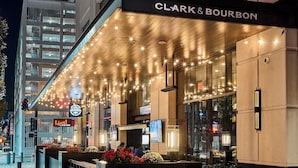 The exterior of the Clark & Bourbon hotel with a well-lit entrance, a marquee sign, and a landscaped planter area.