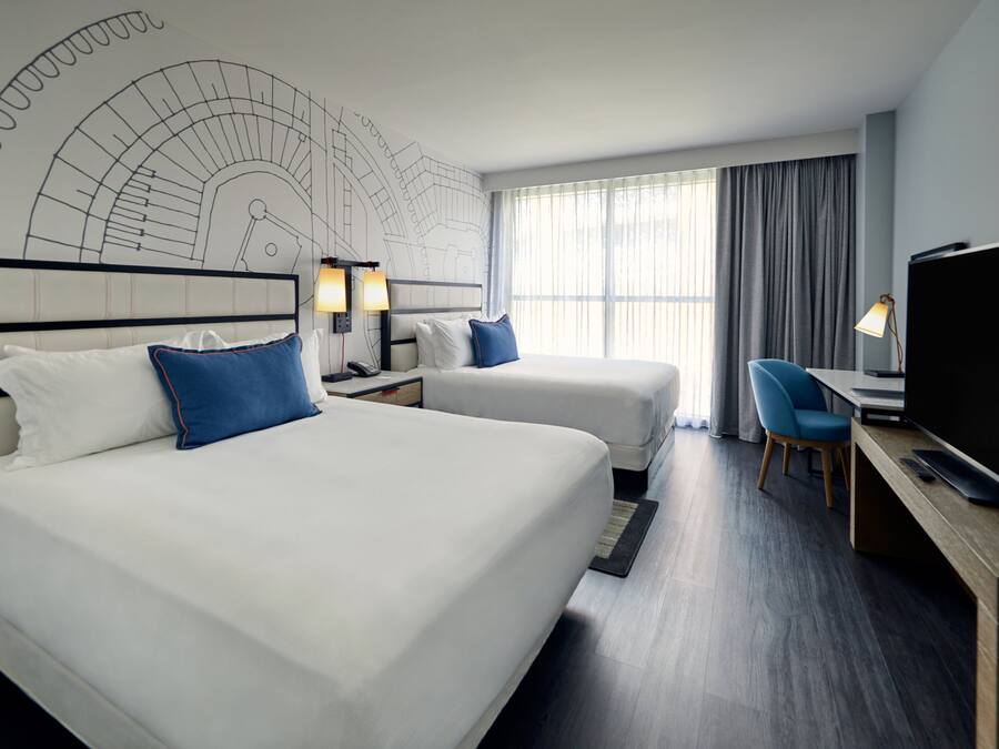 Habitación de hotel con dos camas, un escritorio con computadora, un televisor y un ventanal con cortinas.