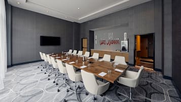 A conference room with a long table, white chairs, a flat-screen TV, and a carpeted floor.