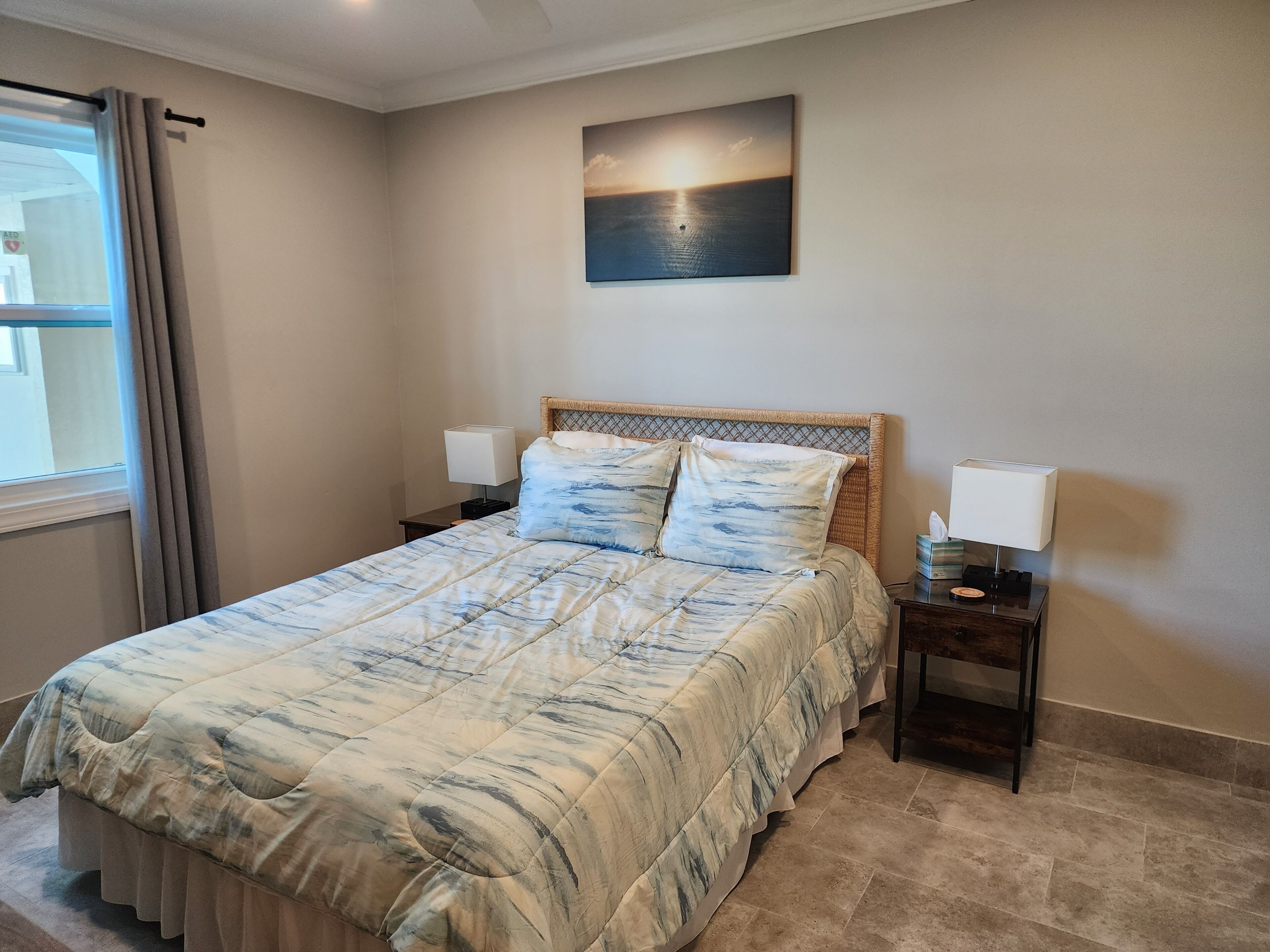 A neatly made bed with a patterned bedspread, two bedside lamps, a nightstand with a tissue box, and a painting of a sunset on the wall.