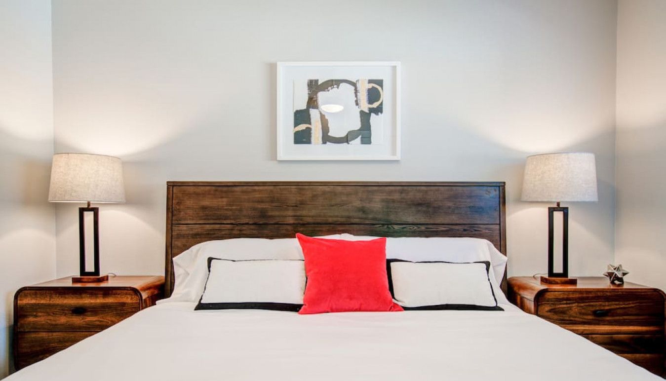 Una cama bien hecha con ropa de cama blanca y negra, una almohada decorativa roja y dos mesitas de noche con lámparas.