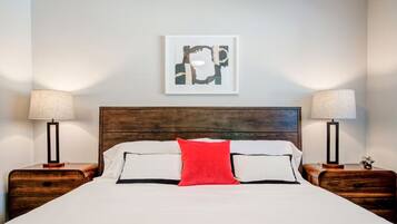 Una cama bien hecha con ropa de cama blanca y negra, una almohada decorativa roja y dos mesitas de noche con lámparas.