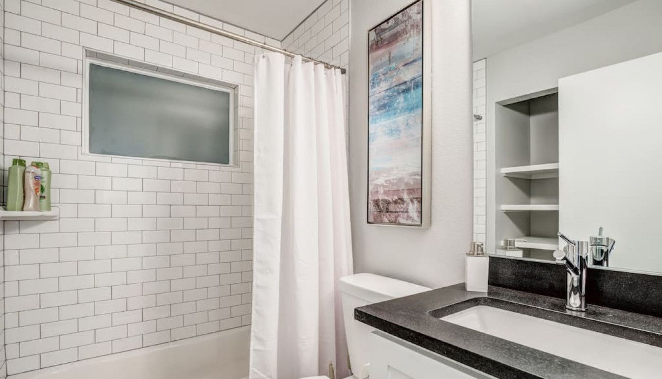Een badkamer met witte metro tegels, een wit toilet, een douche met een witte gordijn, een wastafel met een zwart aanrechtblad en een ingelijst foto aan de muur.