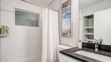 Een badkamer met witte metro tegels, een wit toilet, een douche met een witte gordijn, een wastafel met een zwart aanrechtblad en een ingelijst foto aan de muur.