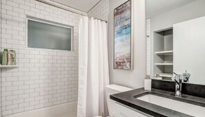 A bathroom with white subway tiles, a white toilet, a shower with a white curtain, a sink with a black countertop, and a framed picture on the wall.