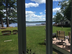 A view through a window of a grassy area with wicker chairs, a wooden deck, and a lake.