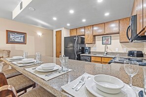 A modern kitchen with granite countertops, wooden cabinets, a stainless steel refrigerator, and a dining area set with plates and glasses. - Walk to Town Lift| Mountain View Balcony | Hot Tub| Managed by Park City Lodging (Park City)