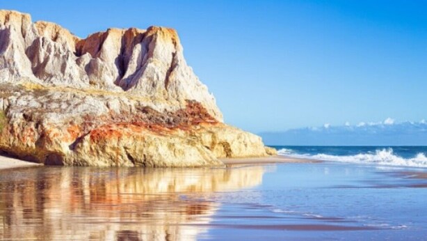 A beach with a rocky cliff and calm water reflecting the sky.