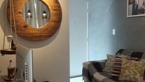 A modern living room with a round wooden mirror, a flat-screen TV, a sofa with patterned cushions, and a checkered rug.
