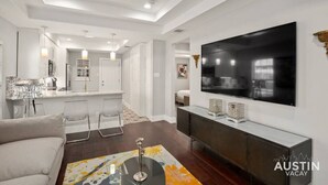 A modern living room with a large flat-screen TV, a dining area with bar stools, and a kitchen in the background. - Olander xxxxxxxxxxxx (Austin)