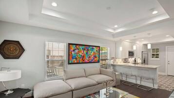 A modern living room with a sofa, a coffee table, and a painting. There is a kitchen area with a bar and a refrigerator.