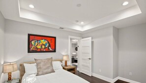 A bedroom with a large bed, a nightstand, a lamp, a door leading to another room, and a colorful painting on the wall.