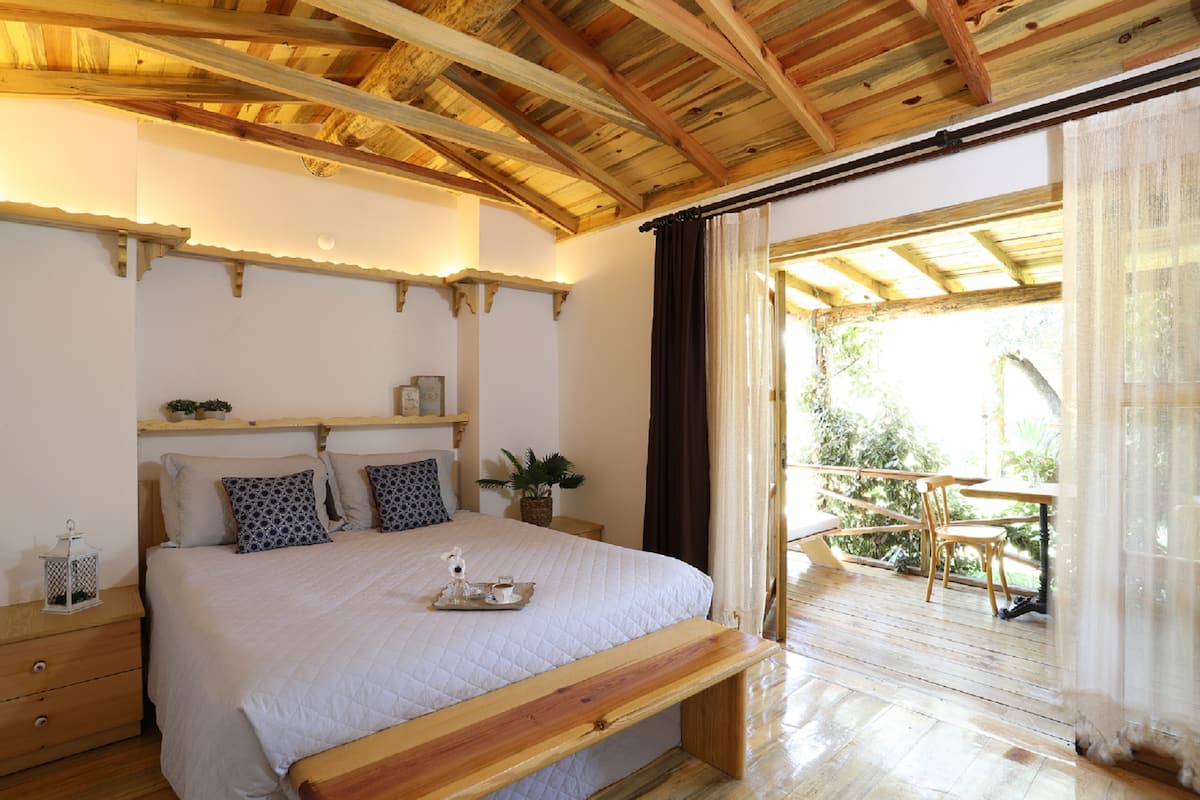 Ein Schlafzimmer mit Holzbalkendecke, einem Bett mit weißer Bettdecke, einem Nachttisch mit Laterne und Blick auf einen Balkon mit Sitzgelegenheiten.