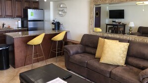 A modern kitchen with dark wooden cabinets, a stainless steel refrigerator, and a granite countertop. There are two yellow bar stools at the kitchen island. - LONG TERM RENTAL ONLY- Oceanfront 1bed/1 bath condo (Daytona Beach Shores)