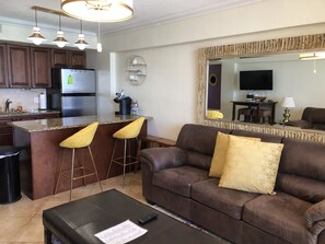 A modern kitchen with dark wooden cabinets, a stainless steel refrigerator, and a granite countertop. There are two yellow bar stools at the kitchen island.