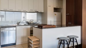 A modern kitchen with white cabinets, stainless steel appliances, a gas stove, and a built-in oven. There are two stools at the island and a wooden rack with dishes. - Mika, 2bdr Fitzroy pad With Secure Parking (Fitzroy)
