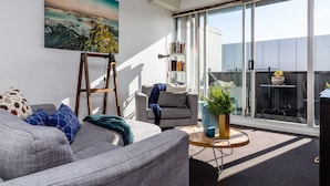 A modern living room with a grey sofa, a wooden ladder, a bookshelf, and a large window with a cityscape view. - Mika, 2bdr Fitzroy pad With Secure Parking (Fitzroy)