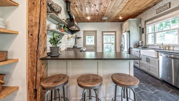 A kitchen with a wooden ceiling, a central island with stools, and a view of the outdoors through large windows. - A REAL Fixer Upper - Giraffe House (Waco)