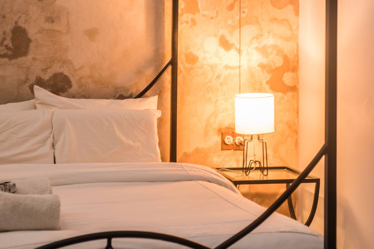 Una cama con ropa de cama blanca y estructura metálica, una mesita de noche con lámpara, y una pared texturizada al fondo.