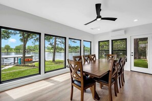 A dining area with a large table and chairs, large windows with a view of a lake and trees, and a ceiling fan. - Wake Zone-Lakefront w/ 1 Boat Lift & Dog Friendly (Kingsland)