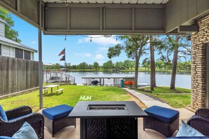Een overdekt terras met rieten meubilair, een vuurkorf en uitzicht op een meer en een park.