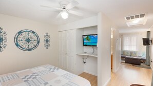 Un dormitorio con cama, ventilador de techo, televisor sobre un soporte y vista a un salón con sofá y ventana.