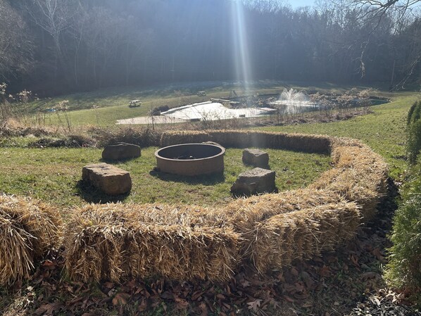 A circular stone fire pit surrounded by hay bales and rocks, with a grassy area and trees in the background. - Family Time at Hills Hocking @ Pine Creek Villas: Creek Front (Rockbridge)