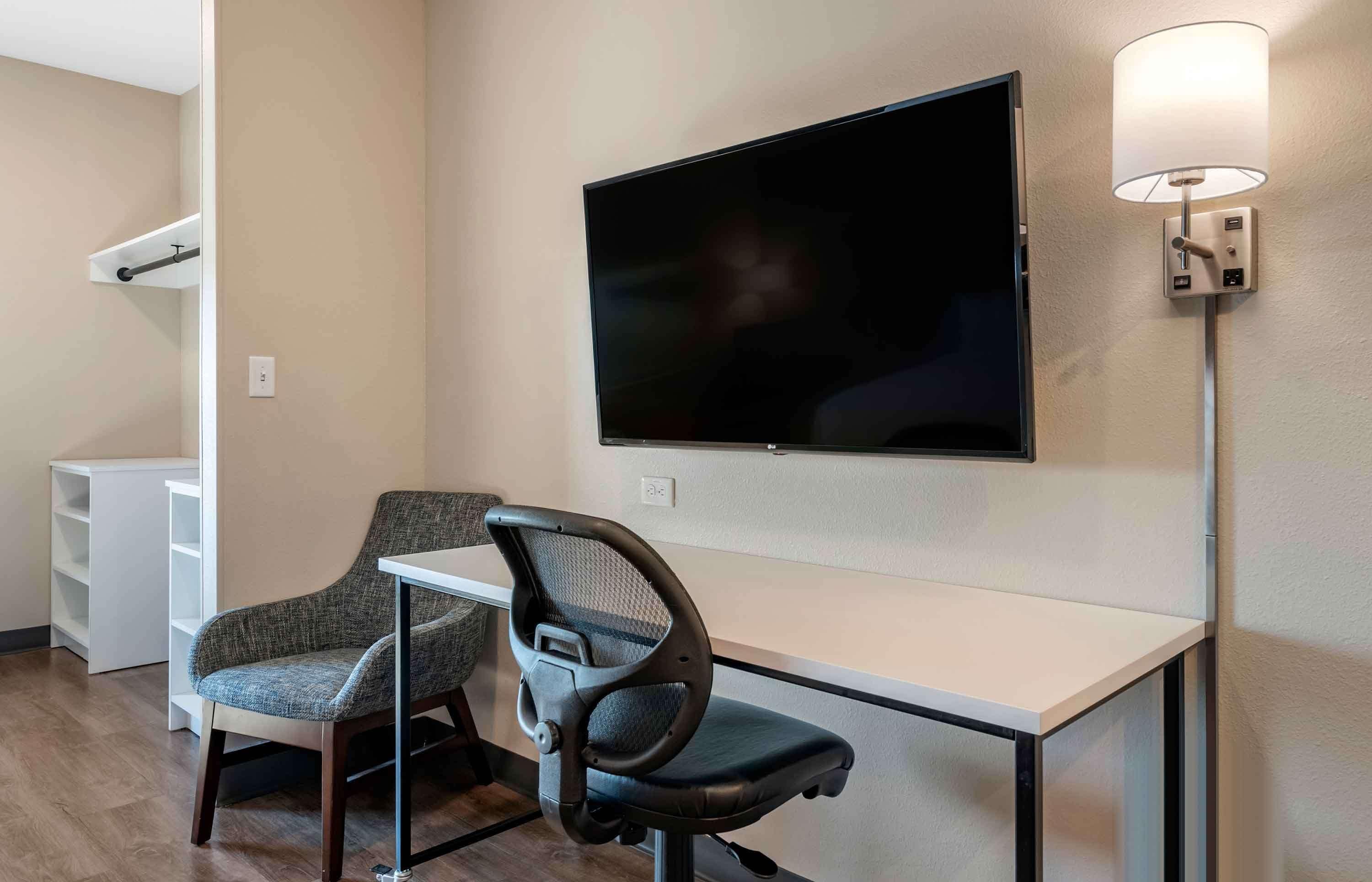 A hotel room with a desk, two chairs, a flat-screen TV mounted on the wall, and a wall-mounted lamp.
