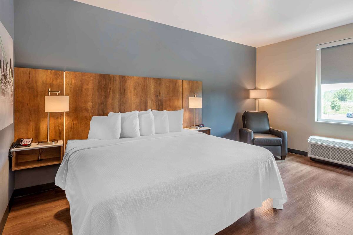 Una habitación de hotel moderna con una cama grande, cabecera de madera, dos mesitas de noche con lámparas, una silla y una ventana con vistas.