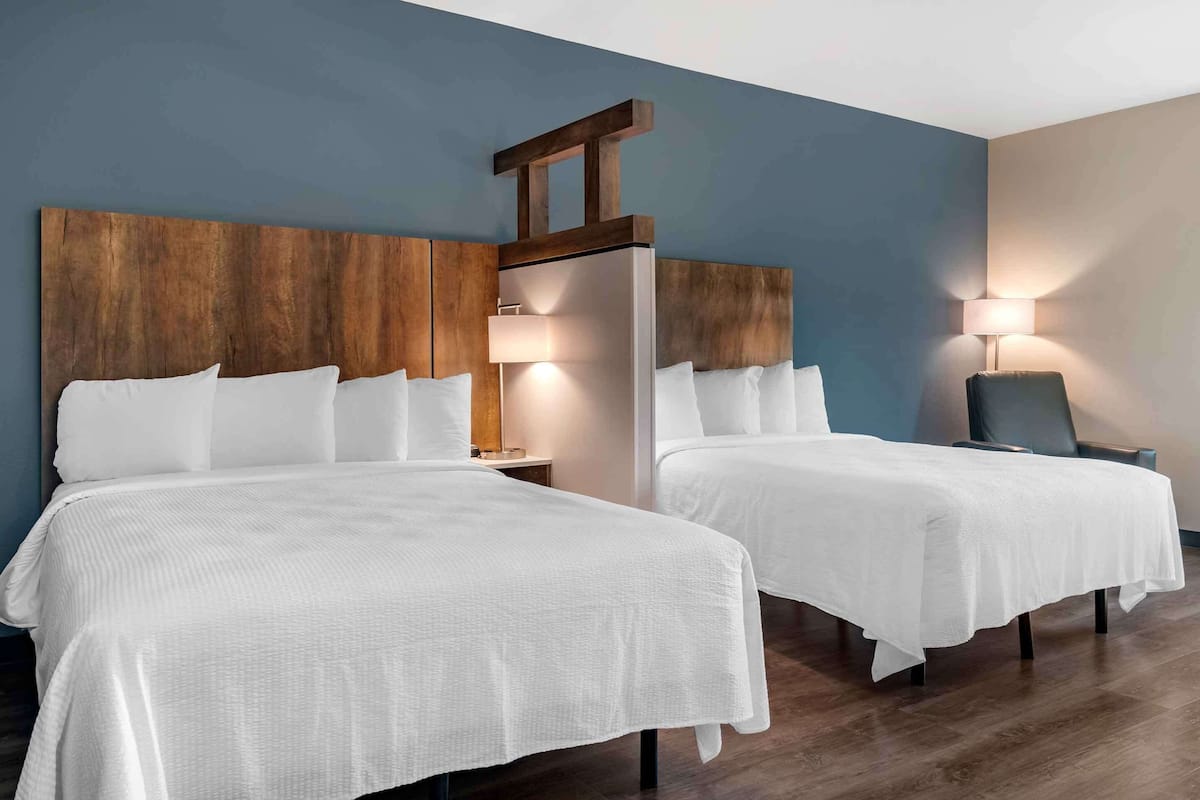 Habitación de hotel con dos camas, cabeceros de madera, ropa de cama blanca, una mesita de noche, una lámpara y una silla.