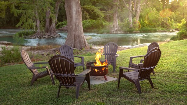 A fire pit with chairs arranged around it in a grassy area near a river, surrounded by trees.