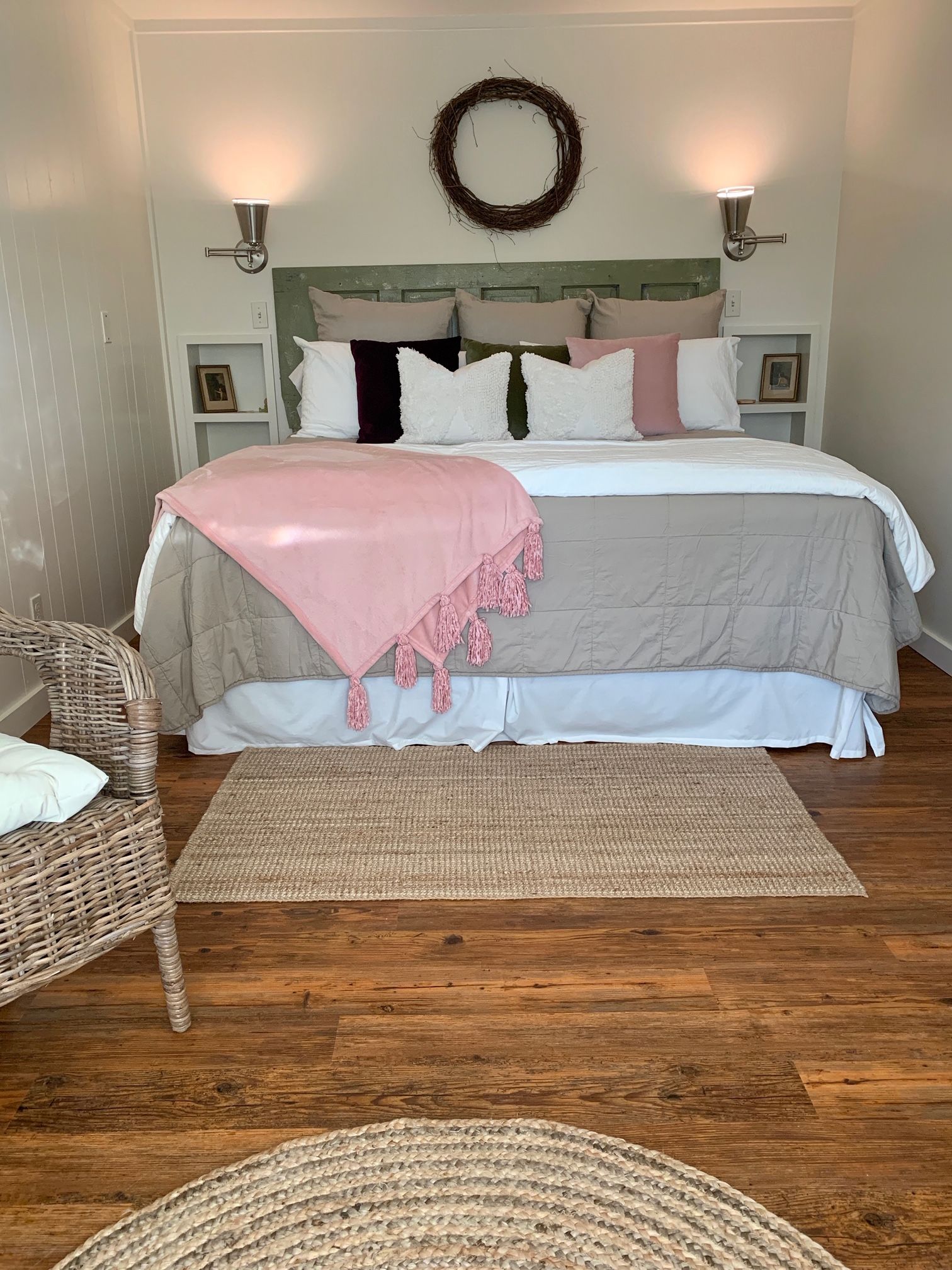 A bedroom with a bed, two bedside tables, a wicker chair, a woven rug, and a wall-mounted wreath.