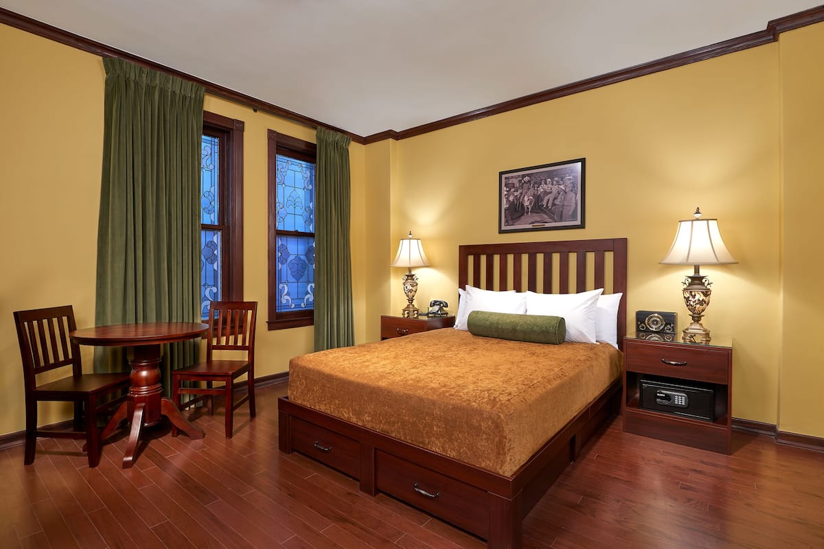 A hotel room with a wooden bed, two bedside tables with lamps, a small table with chairs, and a window with curtains.