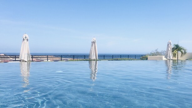 A clear blue infinity pool with three white umbrellas, a view of the ocean, and a fence in the background. - Cabo Private 3bed,3bath Condo + Fober Optic WiFi, Spacious Condo at Quivira Golf (Cabo San Lucas)
