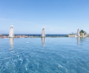 Piscina infinita azul cristalina con tres sombrillas blancas, vista al océano y una cerca al fondo.