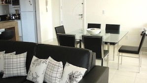 Una sala moderna con un sofá negro, una mesa de comedor de vidrio y una zona de cocina con refrigerador y microondas.