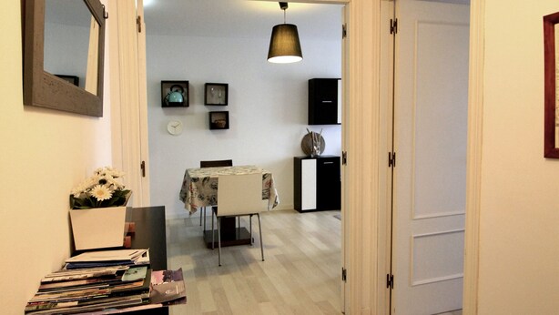 A hallway with a wooden floor, a rug, a mirror, and a shelf with books and a vase of flowers. - Avilés-Salinas V. U.T. 612-AS (salinas/castrillon)