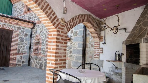Un espace extérieur rustique aménagé avec des murs en pierre et un four en briques, comprenant une table et des chaises placées sous une marquise rose.