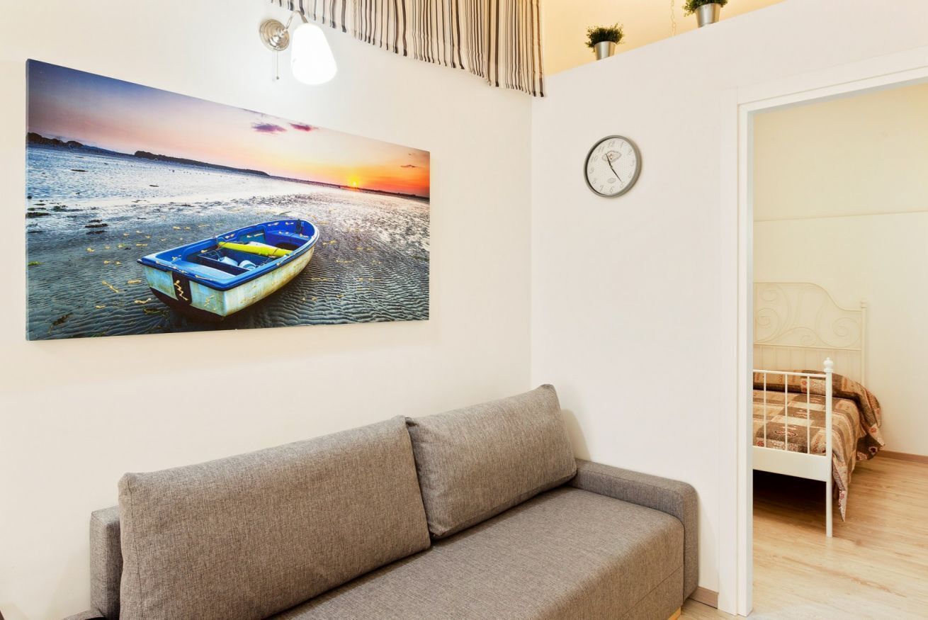 A room with a grey sofa, a bed with a white headboard, a wall clock, and a sunset painting.